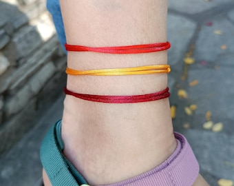 May include: Three thin cord anklets worn around the ankle. The top anklet is bright red, the middle anklet is golden yellow, and the bottom anklet is a deep burgundy red. Each anklet consists of a smooth, slender string material wrapped around.