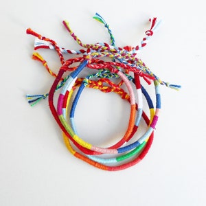 May include: A set of colorful friendship bracelets made with woven thread. The bracelets are in shades of red, blue, green, yellow, and white.