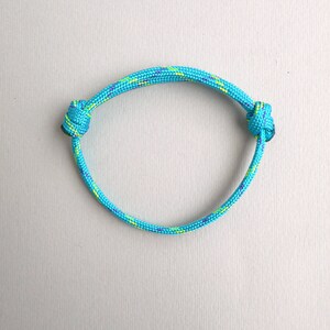 May include: A bright turquoise cord bracelet featuring a woven texture with small flecks of yellow and purple throughout. The bracelet is constructed from a single piece of cord with two adjustable sliding knots on either side of the circular band.