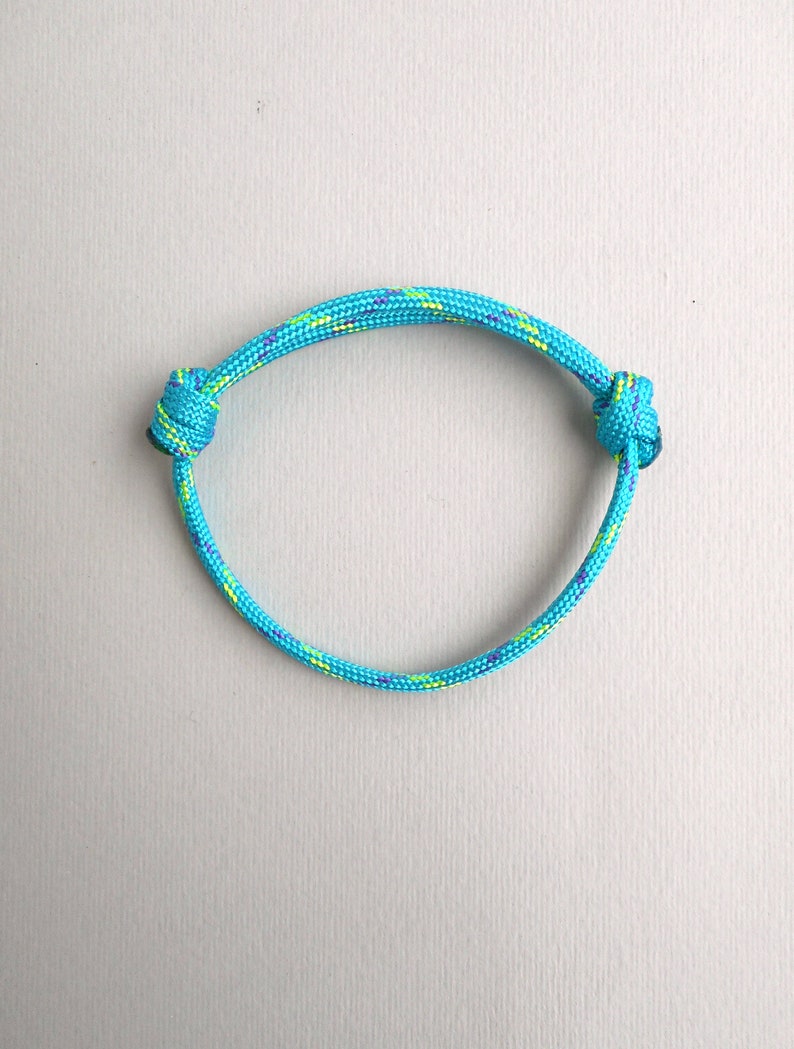 May include: A bright turquoise cord bracelet featuring a woven texture with small flecks of yellow and purple throughout. The bracelet is constructed from a single piece of cord with two adjustable sliding knots on either side of the circular band.