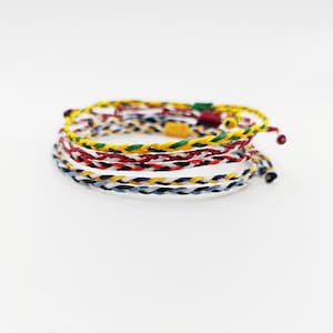 May include: A stack of colorful braided bracelets. The bracelets are in various colors, including yellow, red, white, blue, and green. The bracelets are made of a woven material and are stacked on top of each other.
