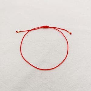 May include: A red string bracelet featuring a sliding knot and gold-coloured beads. The bracelet is shown on a white background. The design is simple and minimalist, suitable for everyday wear.