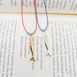 Può includere: Tre collane con ciondoli a forma di pesce. Le collane sono su un libro con del testo. I ciondoli a forma di pesce sono dorati, argentati e blu. Le collane sono su cordini rossi, blu e bianchi.
