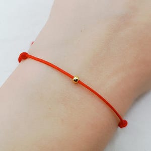May include: A thin red cord bracelet featuring a single small, polished gold-colored spherical bead centered on the string. The bracelet is secured with a simple knot closure, showcasing a minimalist design with a vibrant red nylon thread.
