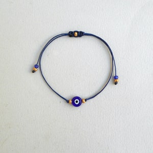 Op de afbeelding: Een donkerblauw koordarmbandje met een centrale ronde kraal in de vorm van een boze oog in blauw, wit en zwart. Het armbandje is versierd met kleine goudkleurige metalen kraaltjes aan weerszijden van de bedel en aan de uiteinden van het koord.