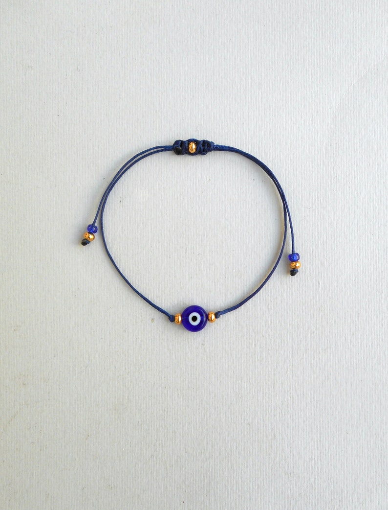 Op de afbeelding: Een donkerblauw koordarmbandje met een centrale ronde kraal in de vorm van een boze oog in blauw, wit en zwart. Het armbandje is versierd met kleine goudkleurige metalen kraaltjes aan weerszijden van de bedel en aan de uiteinden van het koord.