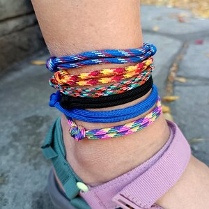 May include: A stack of six cord anklets worn around the ankle. The anklets feature various patterns and colors, including solid blue, solid black, and multi-colored braided designs in shades of yellow, orange, red, green, and pink.