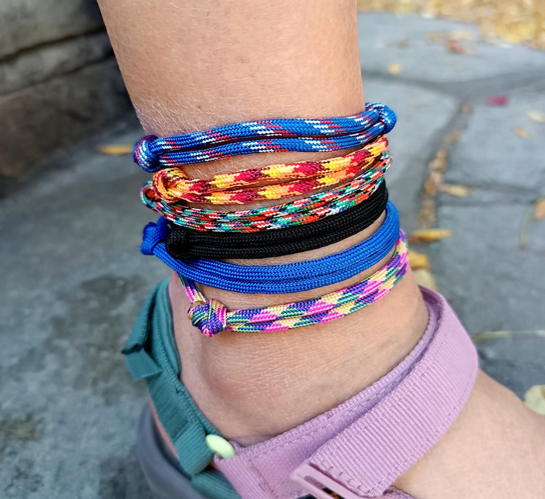 May include: A stack of six cord anklets worn around the ankle. The anklets feature various patterns and colors, including solid blue, solid black, and multi-colored braided designs in shades of yellow, orange, red, green, and pink.