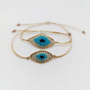 May include: Two handmade macrame bracelets with an evil eye design. The bracelets feature a gold-colored cord and a woven eye with blue, black, and beige colors. The bracelets are adjustable and sit on a white surface.
