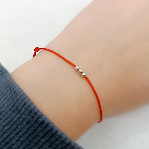 Könnte beinhalten: Ein rotes Schnurarmband mit drei kleinen silbernen Perlen. Das Armband ist am Handgelenk gebunden. Der Hintergrund ist ein heller Hautton und ein dunkelblauer Ärmel.