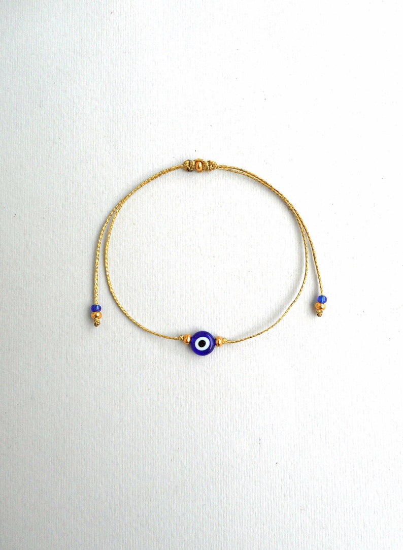 May include: A delicate bracelet featuring a thin gold-colored cord with a central blue and white evil eye bead. The design includes small metallic gold-toned beads flanking the central charm and at the ends of the adjustable cord, which also features tiny blue glass beads.