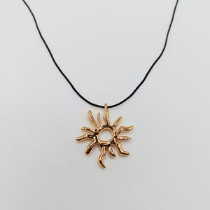 May include: A gold-colored sun pendant necklace with a black cord. The sun pendant has a detailed, textured design with rays extending outward from a central circular opening. The necklace is displayed against a white background.