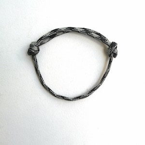 May include: A circular bracelet crafted from a single strand of black, grey, and white camouflage-patterned paracord. The cord is secured with two adjustable sliding knots on opposite sides, allowing the diameter to be expanded or contracted.