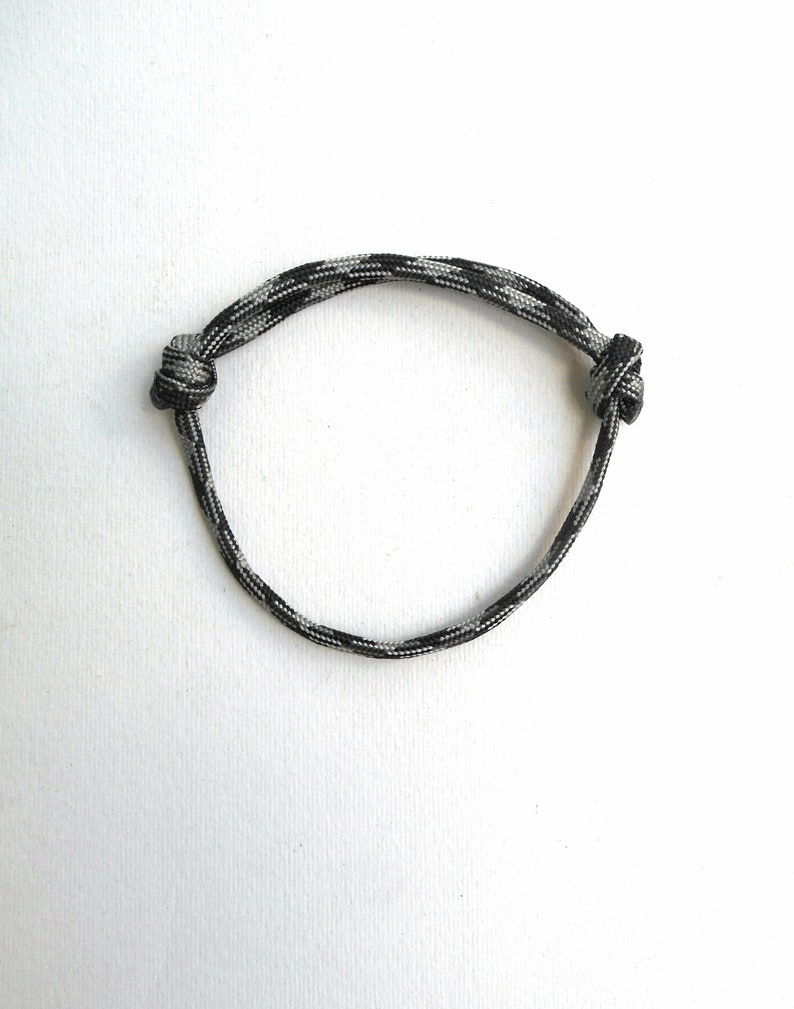 May include: A circular bracelet crafted from a single strand of black, grey, and white camouflage-patterned paracord. The cord is secured with two adjustable sliding knots on opposite sides, allowing the diameter to be expanded or contracted.
