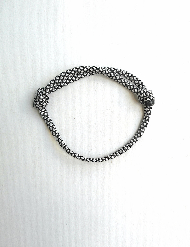 May include: A bracelet constructed from a single length of black and white braided cord. The cord is looped to form a circular shape and secured with two distinct knots on opposite sides, creating a double-strand effect across the top section.