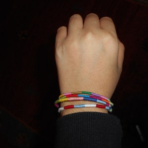 May include: A person's wrist with several colorful bracelets stacked on top of each other. The bracelets are made of woven thread and feature a variety of colors, including red, blue, green, yellow, and pink.