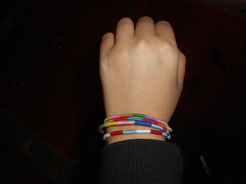 May include: A person's wrist with several colorful bracelets stacked on top of each other. The bracelets are made of woven thread and feature a variety of colors, including red, blue, green, yellow, and pink.