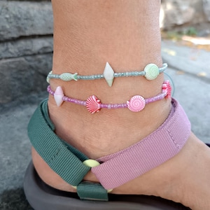 Puede incluir: Dos tobilleras de cuentas con pequeños dijes de temática marina. La tobillera superior tiene cuentas azul claro con una concha blanca y una concha verde pálido. La tobillera inferior tiene cuentas moradas con una concha rosa y una concha a rayas.