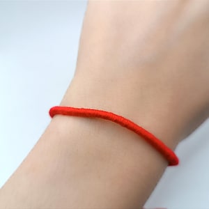 Könnte beinhalten: Ein schlichtes, dünnes Armband aus leuchtend roter Kordel, das um das Handgelenk getragen wird. Das Armband besteht aus einem einzelnen, glatten, durchgehenden Strang aus rotem Material, der das Handgelenk umschließt.