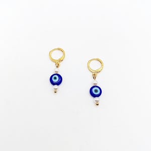 Op de afbeelding: Gouden creolen met een blauwe kwade oog bedel en witte parels.