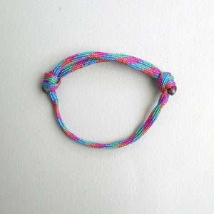 May include: A circular bracelet constructed from a single length of braided cord featuring a vibrant, multi-colored pattern of pink, turquoise, and yellow threads. The cord is secured on both sides by two distinct, tightly tied overhand knots.