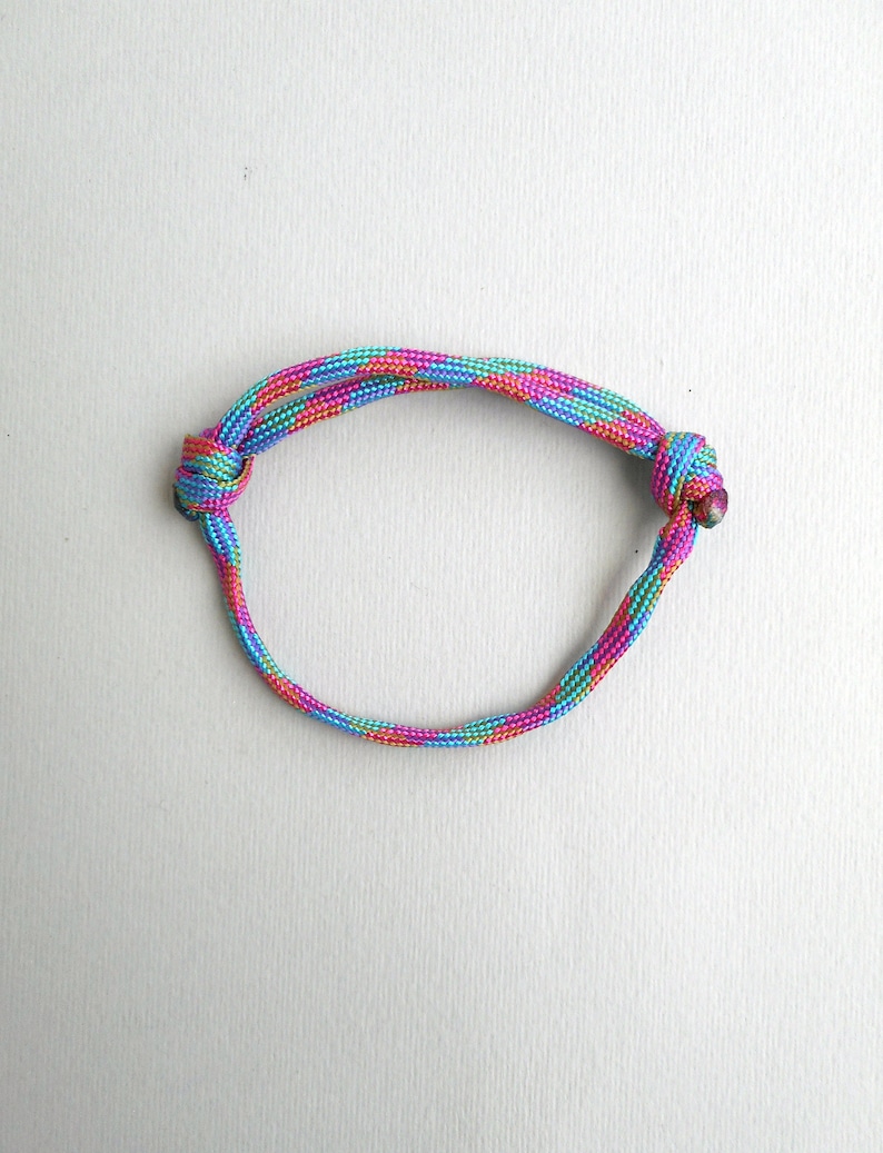 May include: A circular bracelet constructed from a single length of braided cord featuring a vibrant, multi-colored pattern of pink, turquoise, and yellow threads. The cord is secured on both sides by two distinct, tightly tied overhand knots.
