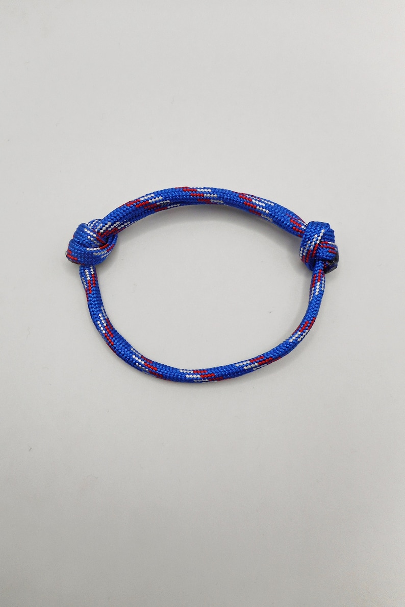 May include: A circular bracelet crafted from blue paracord featuring a woven pattern of small white and red flecks. The cord is tied with two distinct knots on opposite sides, creating a continuous loop design with a textured, braided surface.
