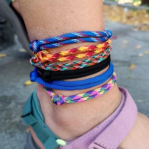 May include: A stack of five cord anklets worn around the ankle. The anklets feature various designs including a blue and white speckled cord, a multi-colored orange and yellow pattern, a green and red mix, a solid black cord, and a pink and blue pattern.