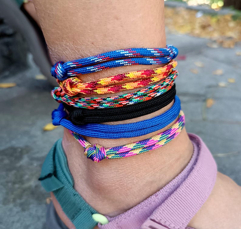 May include: A stack of five cord anklets worn around the ankle. The anklets feature various designs including a blue and white speckled cord, a multi-colored orange and yellow pattern, a green and red mix, a solid black cord, and a pink and blue pattern.