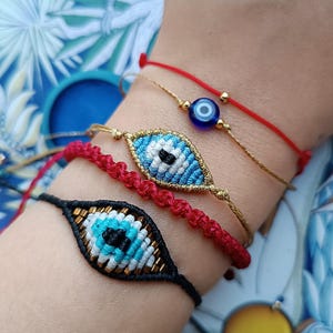 Op de afbeelding: Een set van vier armbanden met twee geweven boze oog bedels, een met een zwart koord en een met een goudkleurig koord, naast een dun rood koordarmbandje met een kleine blauwe glaskraal en een rood gevlochten koordarmbandje met gouden accenten.