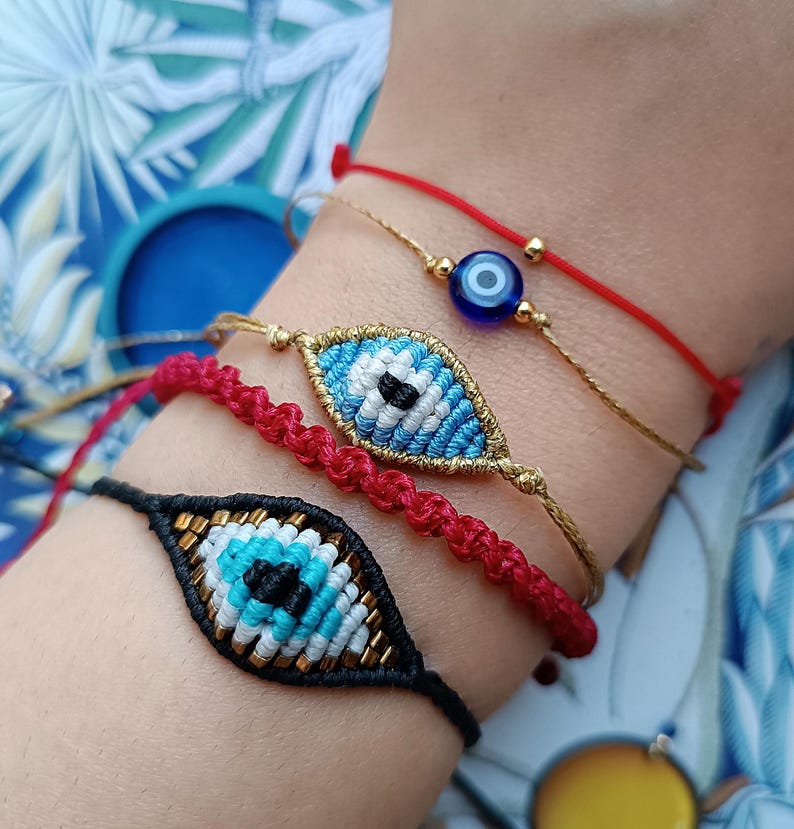 Op de afbeelding: Een set van vier armbanden met twee geweven boze oog bedels, een met een zwart koord en een met een goudkleurig koord, naast een dun rood koordarmbandje met een kleine blauwe glaskraal en een rood gevlochten koordarmbandje met gouden accenten.
