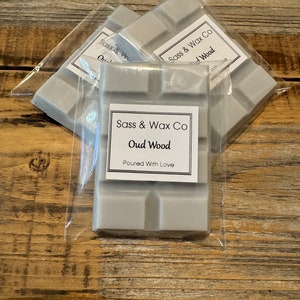 May include: Three wax melts in clear plastic packaging. The wax melts are grey and have the text "Sass & Wax Co Oud Wood Poured With Love" printed on them.