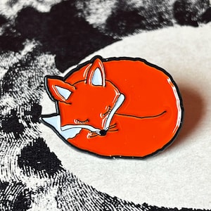 Könnte beinhalten: Ein Emaille-Pin mit einem schlafenden Fuchs. Der Pin zeigt einen zusammengerollten Fuchs in Orange, Weiß und Schwarz. Der Fuchs ist schwarz umrandet, mit weißen Brust- und Ohrendetails. Ein charmantes Accessoire.