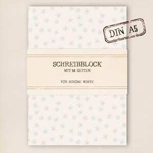 Könnte beinhalten: Ein weißer Schreibblock mit einem blauen Schneeflockenmuster. Der Block hat ein braunes Etikett mit der Aufschrift "Schreibblock mit 50 Seiten" und "Für schöne Worte". Das Etikett hat auch einen Stempel mit der Aufschrift "DIN A5".