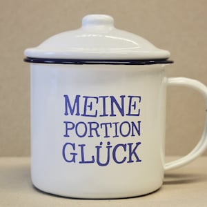 Könnte beinhalten: Weiße Email Tasse mit blauem Deckel und Henkel. Die Tasse hat den Text "MEINE PORTION GLÜCK" in blau aufgedruckt.
