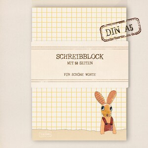 Könnte beinhalten: Ein gelber und weißer kariertes Notizbuch mit einer braunen Hasenillustration. Das Notizbuch hat ein braunes Etikett mit der Aufschrift "SCHREIBBLOCK MIT 50 SEITEN FUR SCHONE WORTE" und einen braunen Stempel mit der Aufschrift "DIN A5".