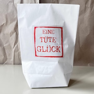 Puede incluir: Una bolsa de papel blanca con una etiqueta roja. La etiqueta presenta el texto alemán "EINE TÜTE GLÜCK" en rojo, con una fuente manuscrita. La bolsa está doblada en la parte inferior y se mantiene de pie.