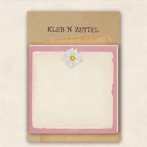 Könnte beinhalten: Notizblock mit rosa Rand und Gänseblümchen-Illustration. Der Notizblock liegt auf einer braunen Papierunterlage. Der Text "KLEB 'N ZETTEL" ist oben auf dem braunen Papier aufgedruckt.