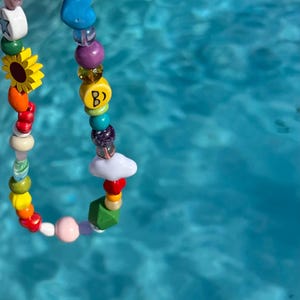 May include: A colorful beaded necklace with a variety of shapes and colors. The necklace includes a sunflower, a star, a heart, and a bead with the letter "B". The beads are strung on a clear cord.