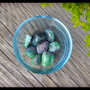 May include: A clear glass bowl filled with polished ruby zoisite stones. The stones are a mix of green and pink, with a speckled appearance. The bowl sits on a weathered wooden surface, with green foliage in the background.
