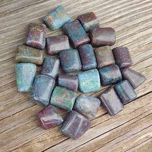 May include: A collection of tumbled stones in shades of teal, burgundy, and green. The stones are rectangular with rounded edges and are arranged on a weathered wooden surface. The stones vary in color and have a polished appearance.