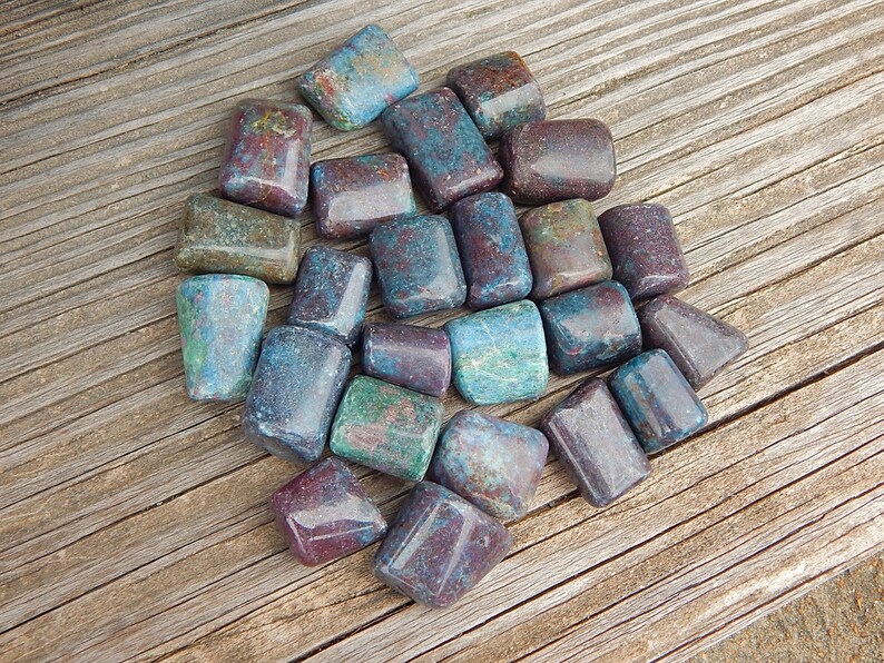 May include: A collection of tumbled stones in shades of teal, burgundy, and green. The stones are rectangular with rounded edges and are arranged on a weathered wooden surface. The stones vary in color and have a polished appearance.