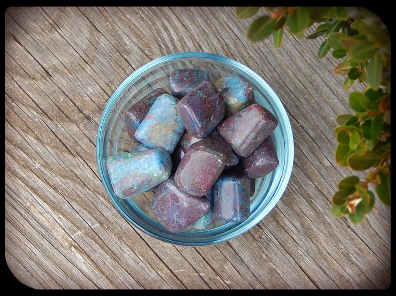 May include: A clear glass bowl filled with tumbled stones. The stones are a mix of colors, including blue, purple, and brown. The bowl sits on a weathered wooden surface, with green foliage in the background.
