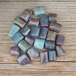 May include: A collection of tumbled stones in shades of blue, green, and burgundy. The stones are rectangular and have a polished, glossy finish. They are scattered on a wooden surface, showcasing their natural colors and textures.