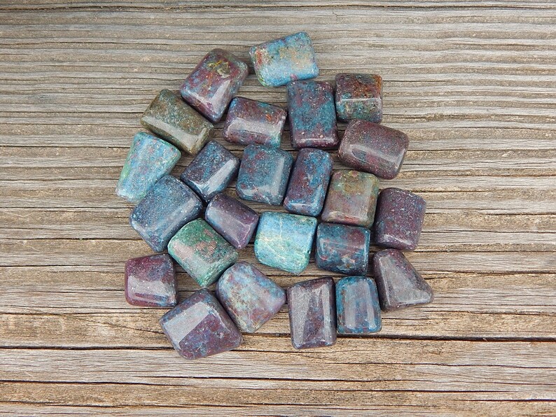 May include: A collection of tumbled stones in shades of blue, green, and burgundy. The stones are rectangular and have a polished, glossy finish. They are scattered on a wooden surface, showcasing their natural colors and textures.
