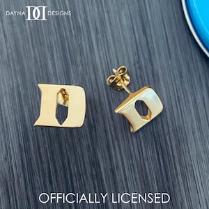 May include: Gold-colored stud earrings in the shape of the letter "D". The earrings are displayed on a dark gray surface. The text "OFFICIALLY LICENSED" is at the bottom of the image.