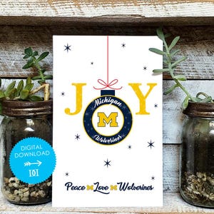 May include: A white card with the word "JOY" in yellow letters. A navy blue and yellow ornament with "Michigan Wolverines" text hangs from a red string. The phrase "Peace, Love, Wolverines" is at the bottom.