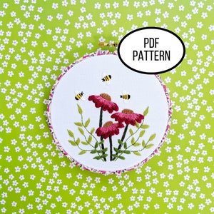 May include: Embroidered hoop art featuring pink flowers and bees. The hoop is white with a pink and white floral trim. The text "PDF PATTERN" is visible in a white oval.