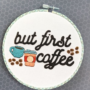 Puede incluir: Arte de bordado en aro con el texto "but first coffee" en fuente cursiva negra. El dise&ntilde;o incluye una taza de caf&eacute; azul y un vaso de caf&eacute; para llevar marr&oacute;n con tapa roja y un c&iacute;rculo blanco en el lateral. Tambi&eacute;n hay granos de caf&eacute; marrones esparcidos alrededor del dise&ntilde;o.