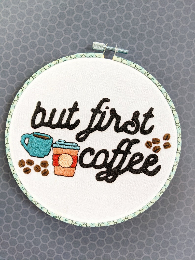 Puede incluir: Arte de bordado en aro con el texto "but first coffee" en fuente cursiva negra. El dise&ntilde;o incluye una taza de caf&eacute; azul y un vaso de caf&eacute; para llevar marr&oacute;n con tapa roja y un c&iacute;rculo blanco en el lateral. Tambi&eacute;n hay granos de caf&eacute; marrones esparcidos alrededor del dise&ntilde;o.